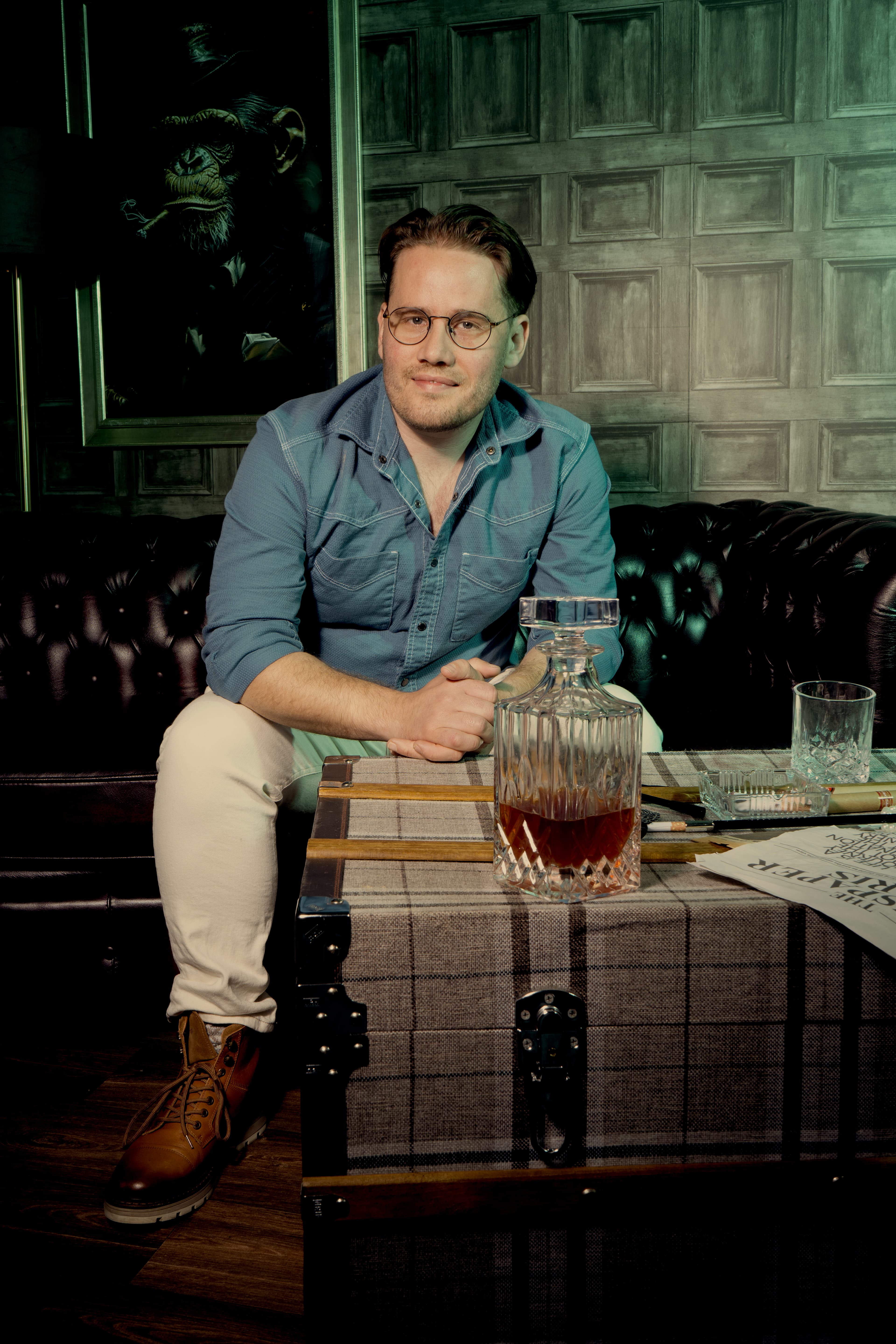 Man in glasses sits on a leather sofa behind a trunk with a whiskey decanter.